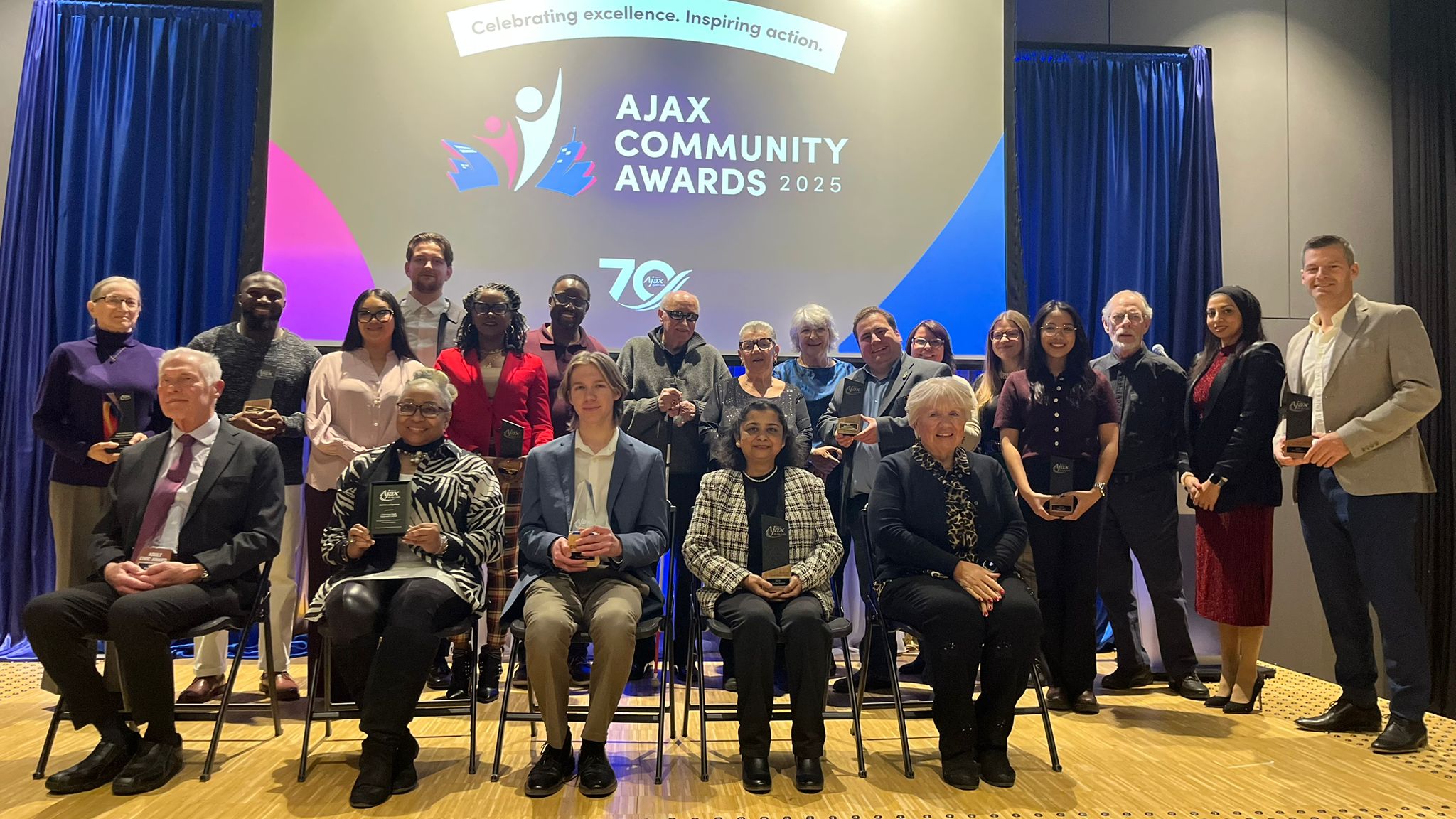 A group of people pose on stage holding awards at the Ajax Community Awards 2025 event.