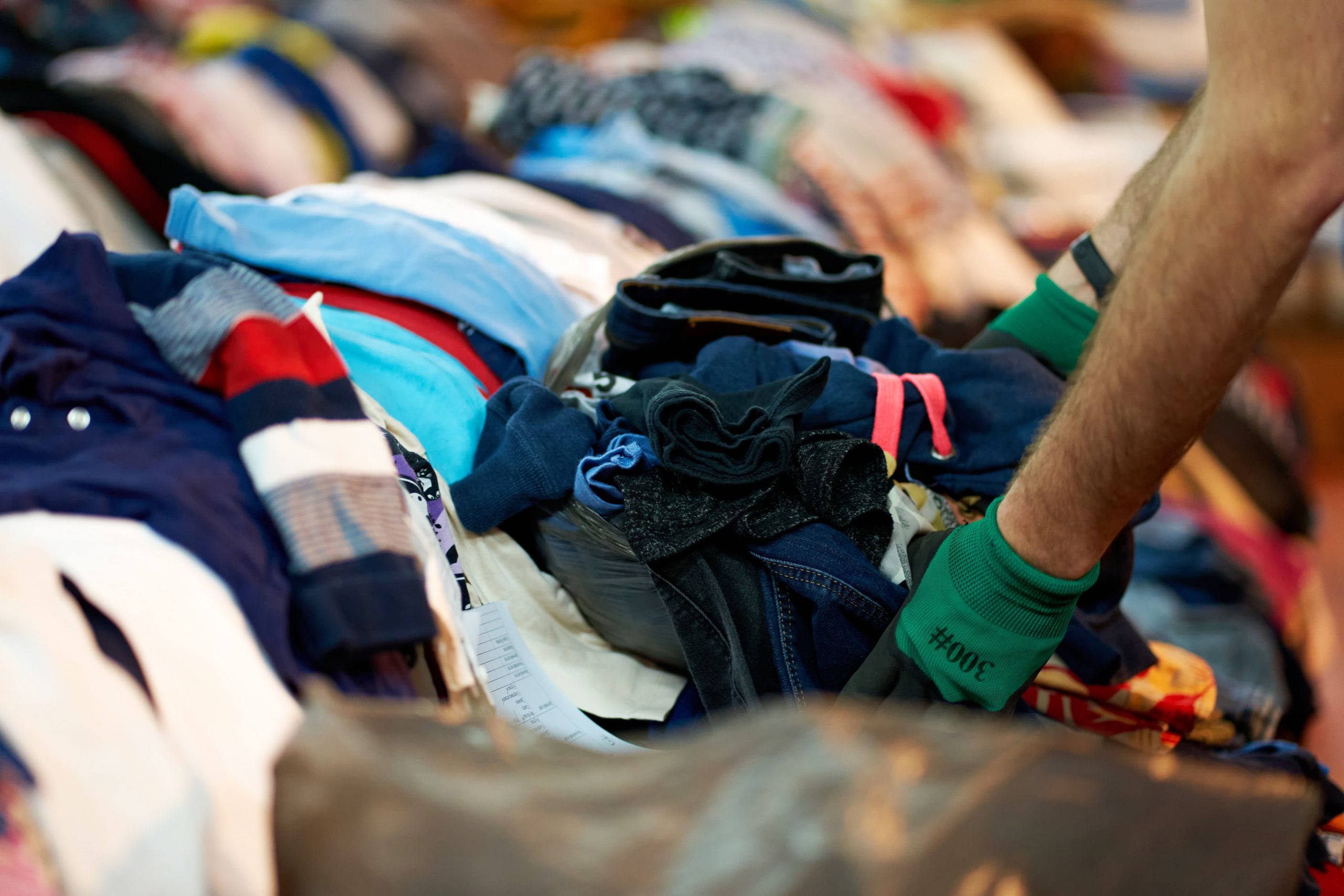 Person wearing gloves sorting through piles of assorted folded clothes on a table.