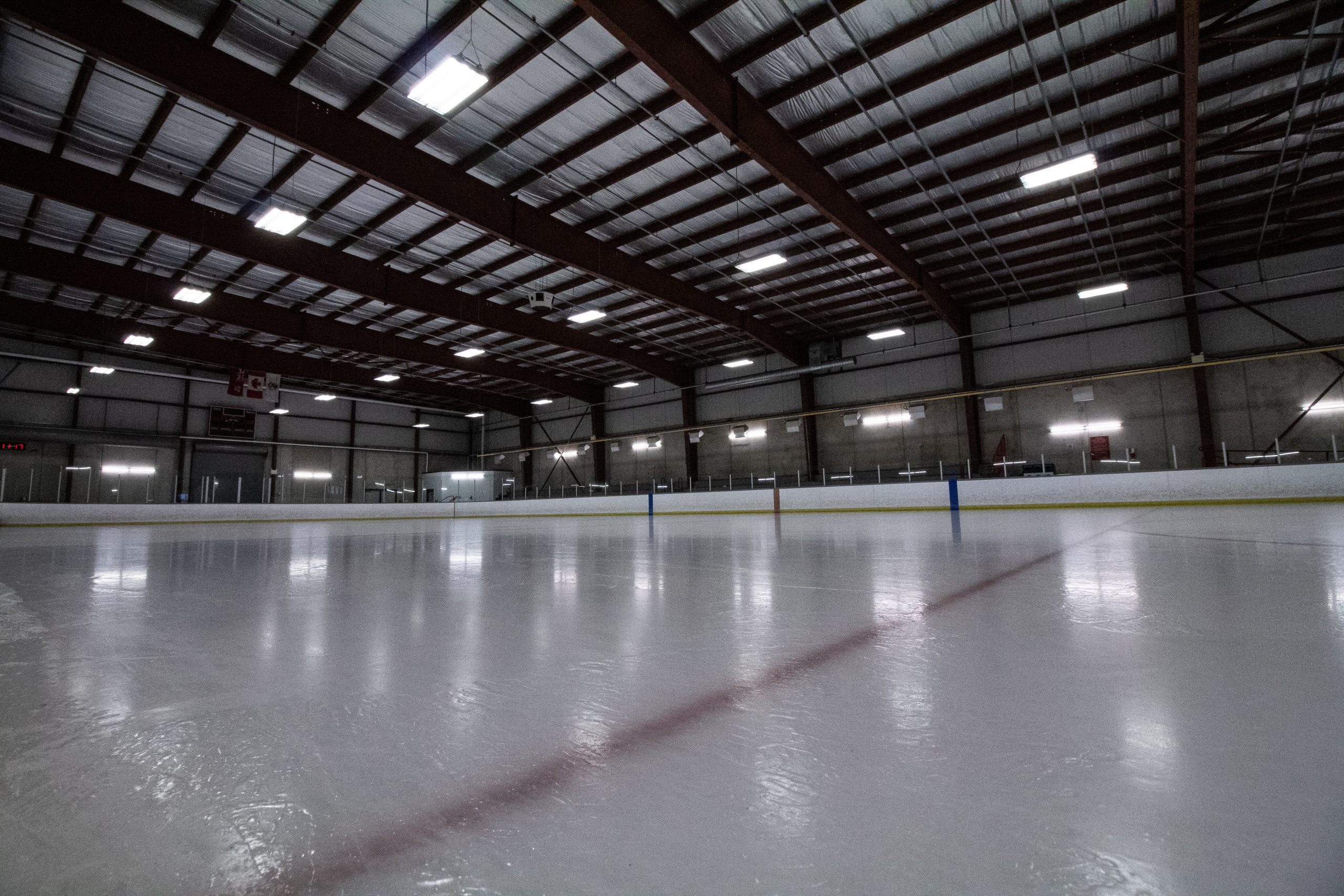 Empty indoor ice rink with visible red and blue lines, surrounded by low barriers and overhead lights.