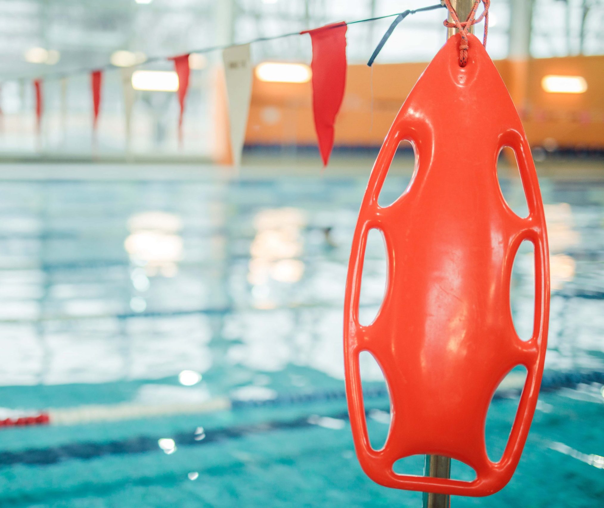 Red lifeguard rescue tube hanging by an indoor swimming pool with lane dividers and flags in the background.