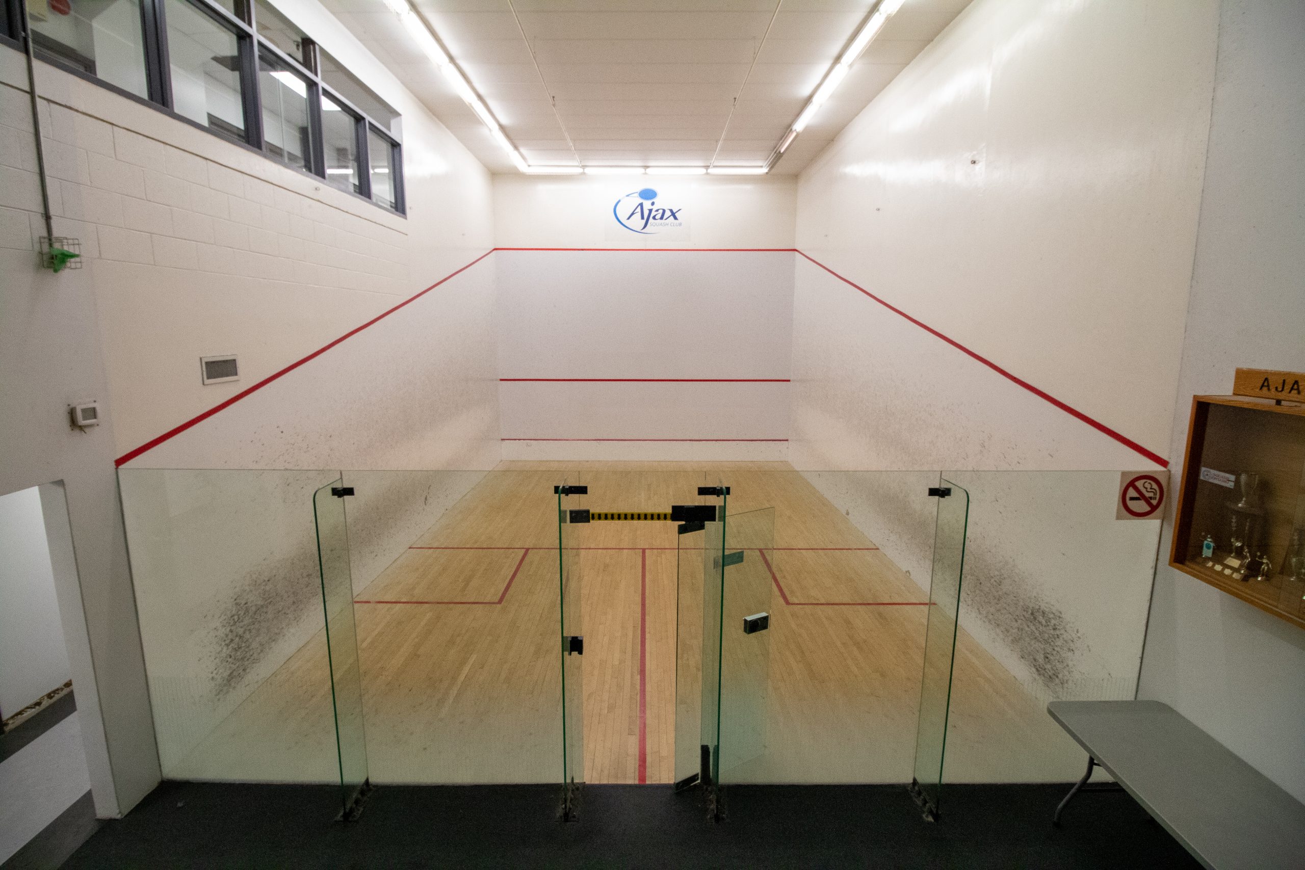Empty indoor squash court with glass doors, wooden floor, white walls, and the word "Ajax" above the front wall.