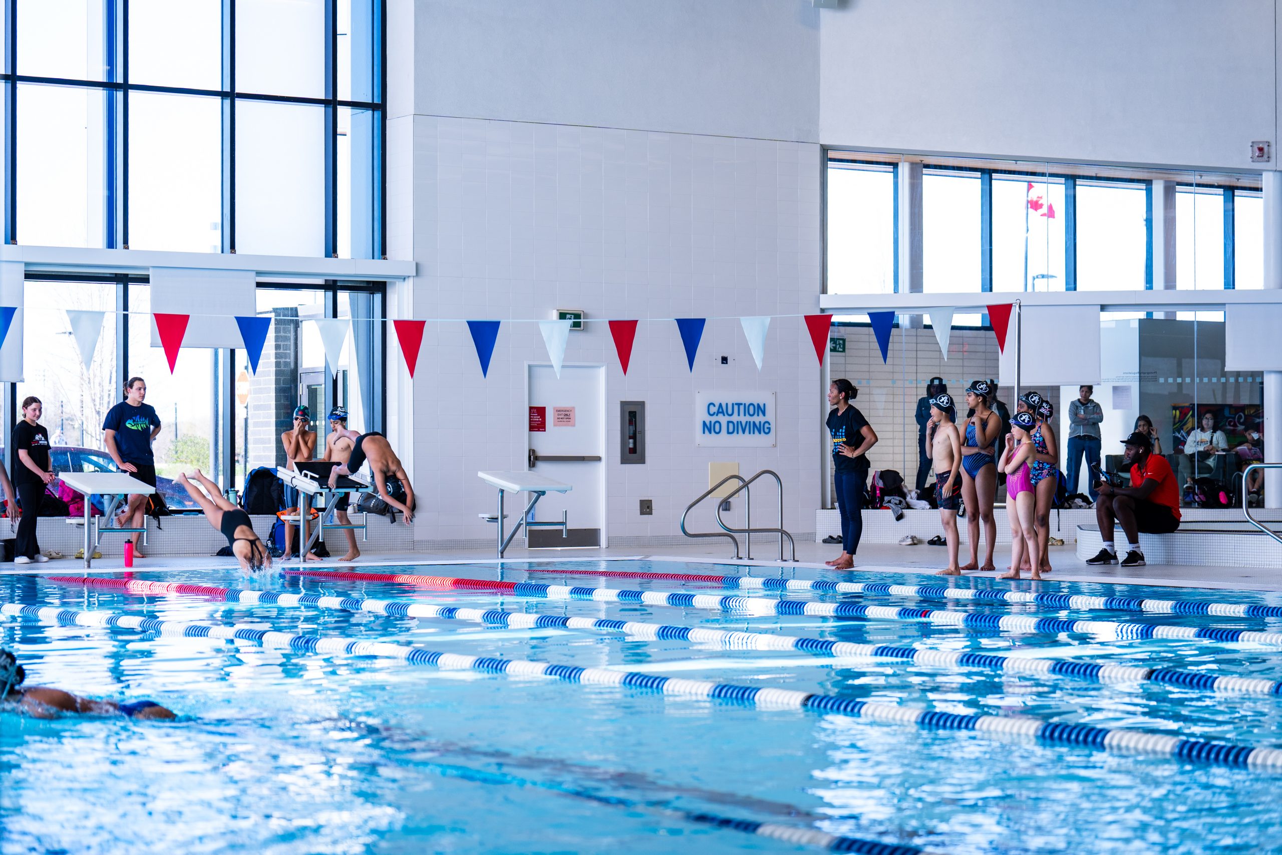 Swimmers prepare to dive into a pool as others watch from the poolside at an indoor swimming facility.