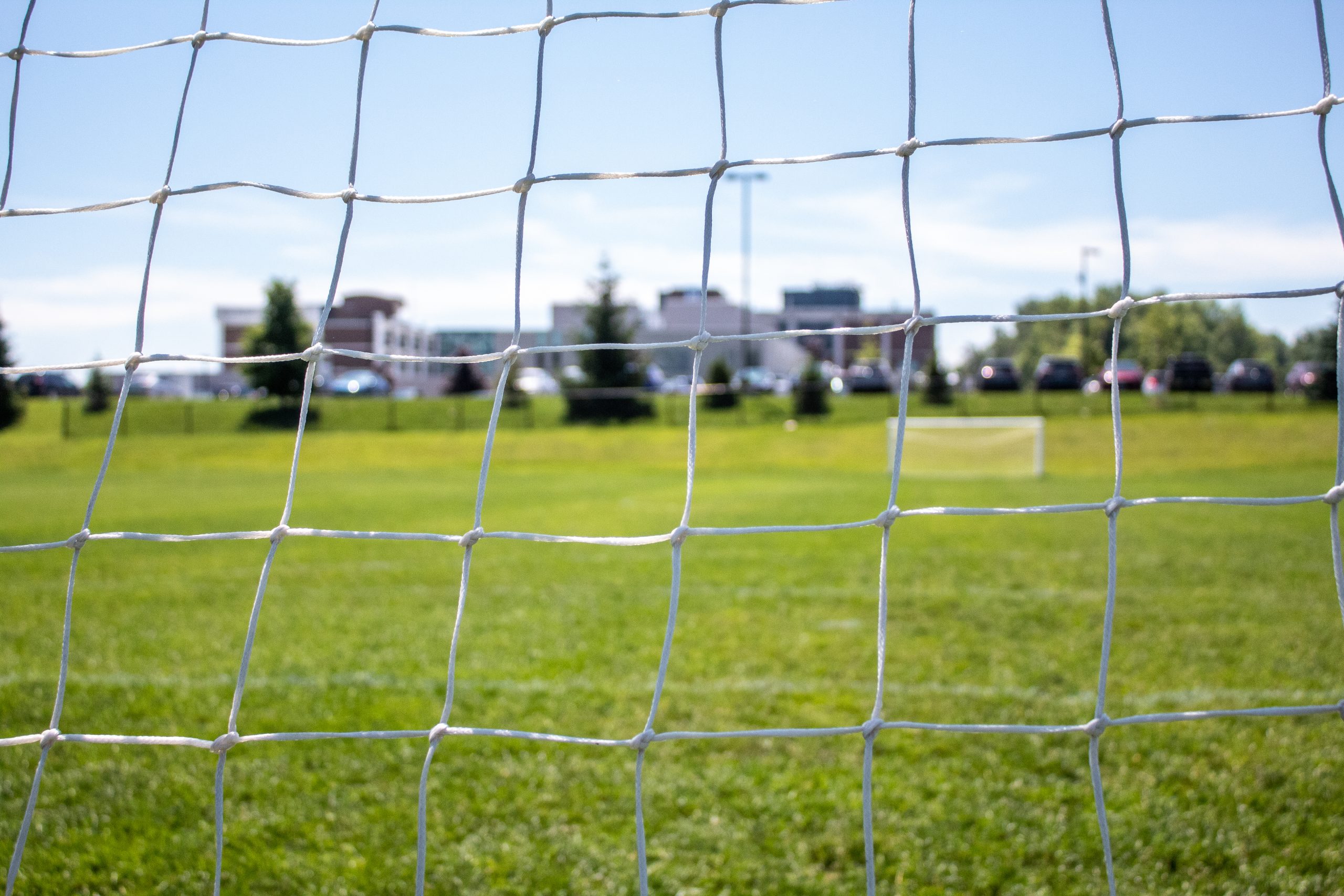 A close-up of a football net with a grassy pitch, goalpost, cars, and buildings in the background under a blue sky.