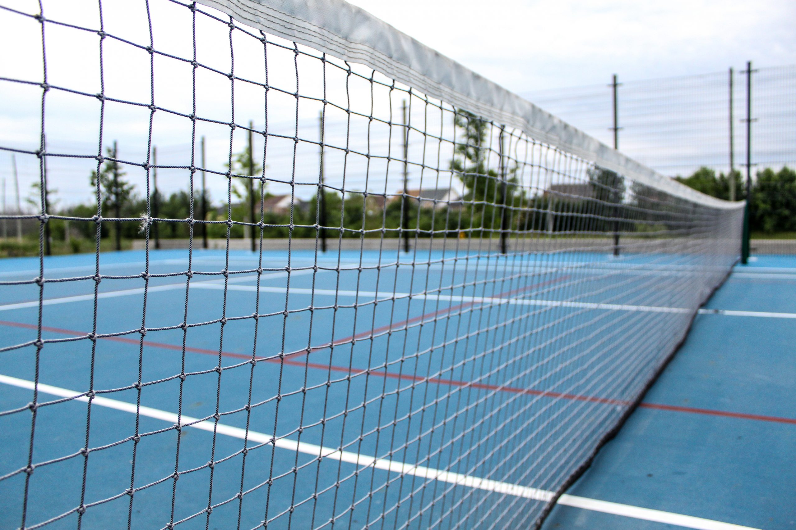 Close-up view of a tennis net on an empty outdoor blue tennis court with trees and fences in the background.