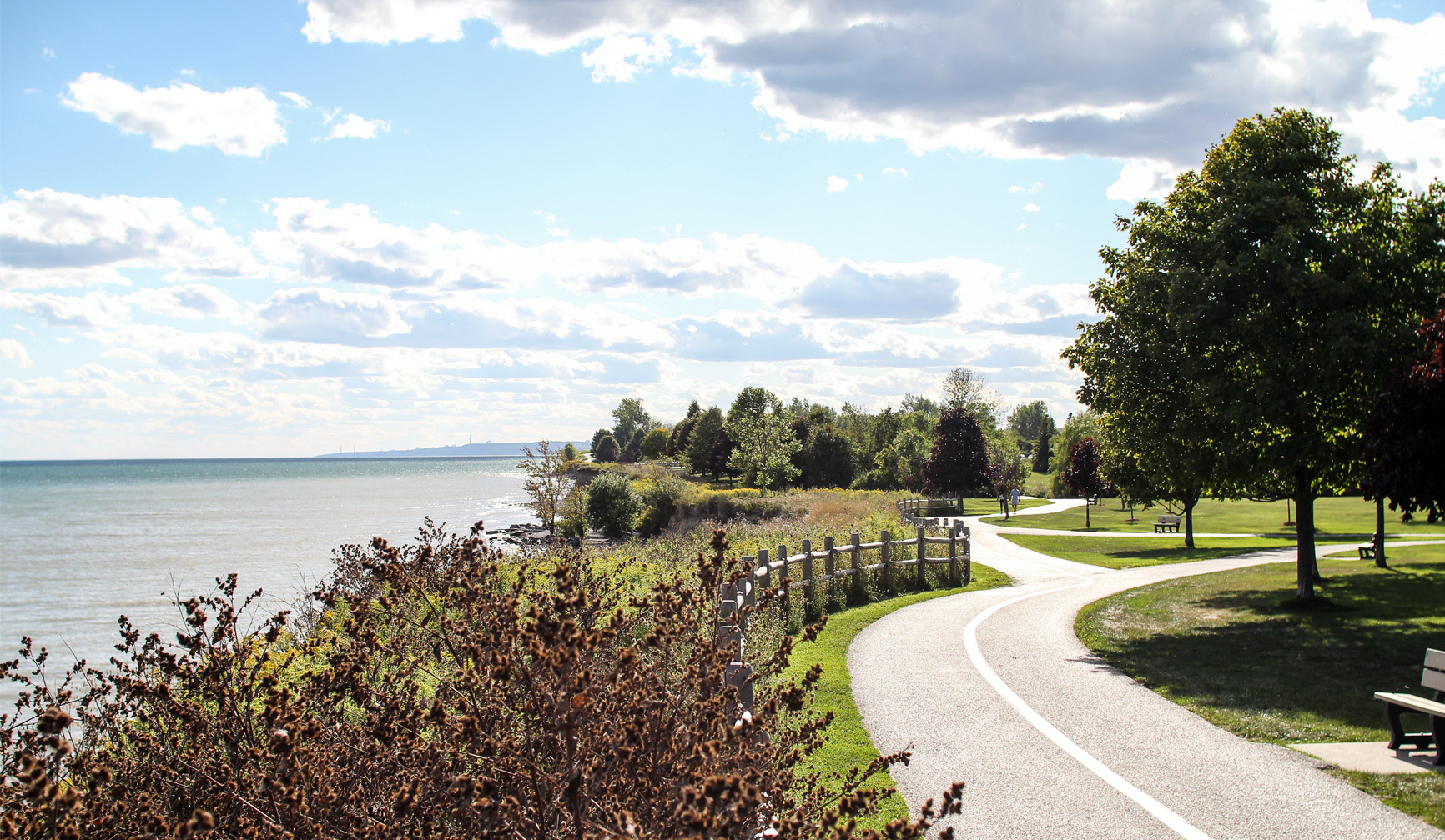 Paved walking path curves along a lakeshore with trees, grass, benches, and partly cloudy sky.