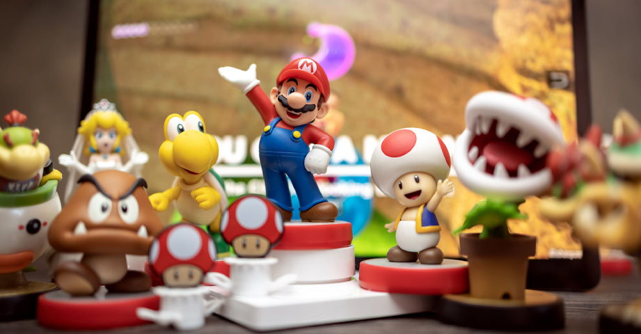 Assorted Super Mario character figurines are arranged in front of a blurred video game screen background.