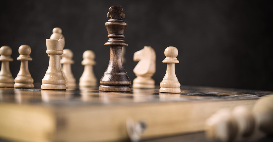 Close-up of chess pieces on a wooden chessboard, with the black king surrounded by white pieces.