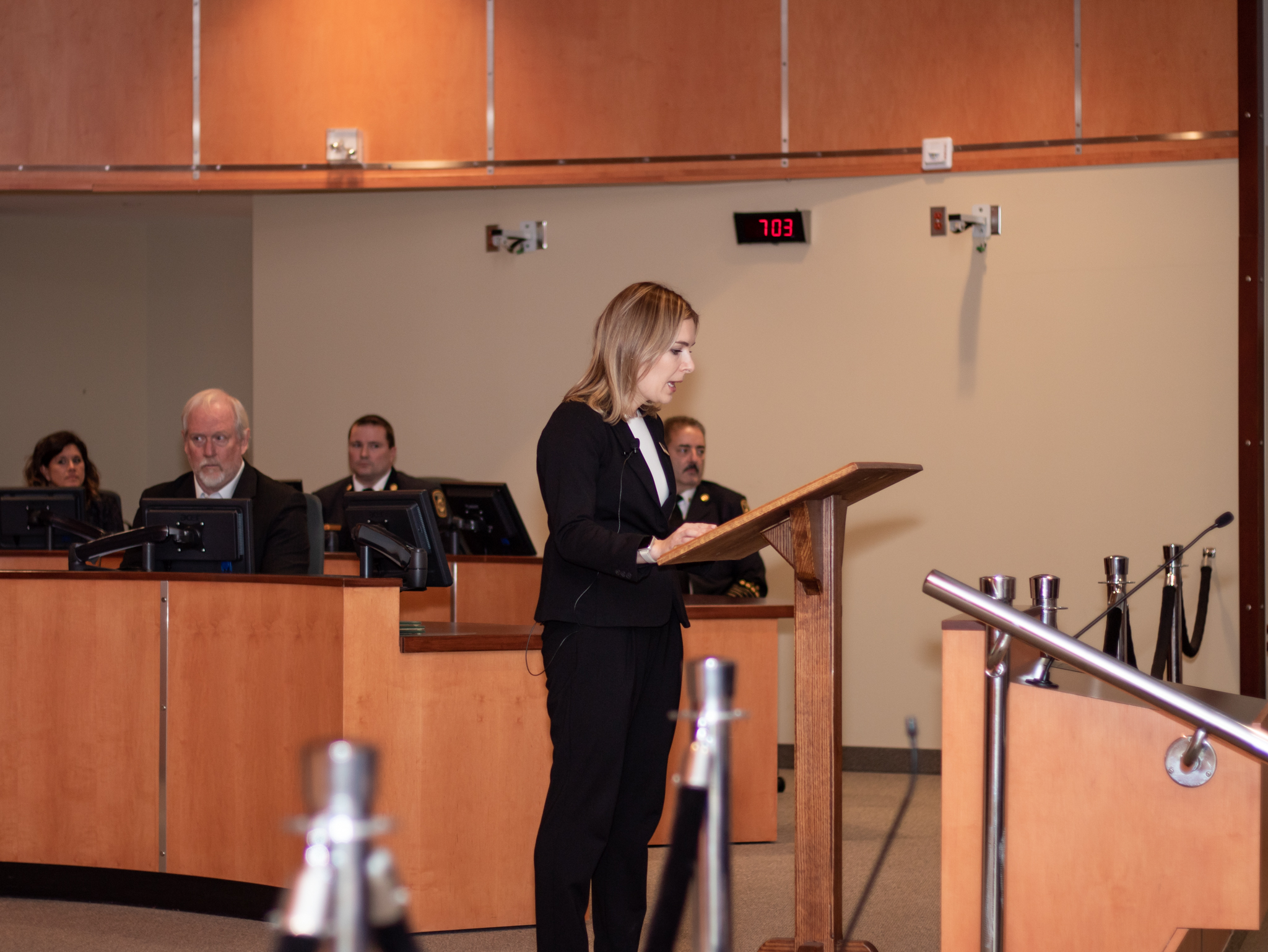 A woman in a suit speaks at a lectern in a formal meeting room with several people seated behind her.