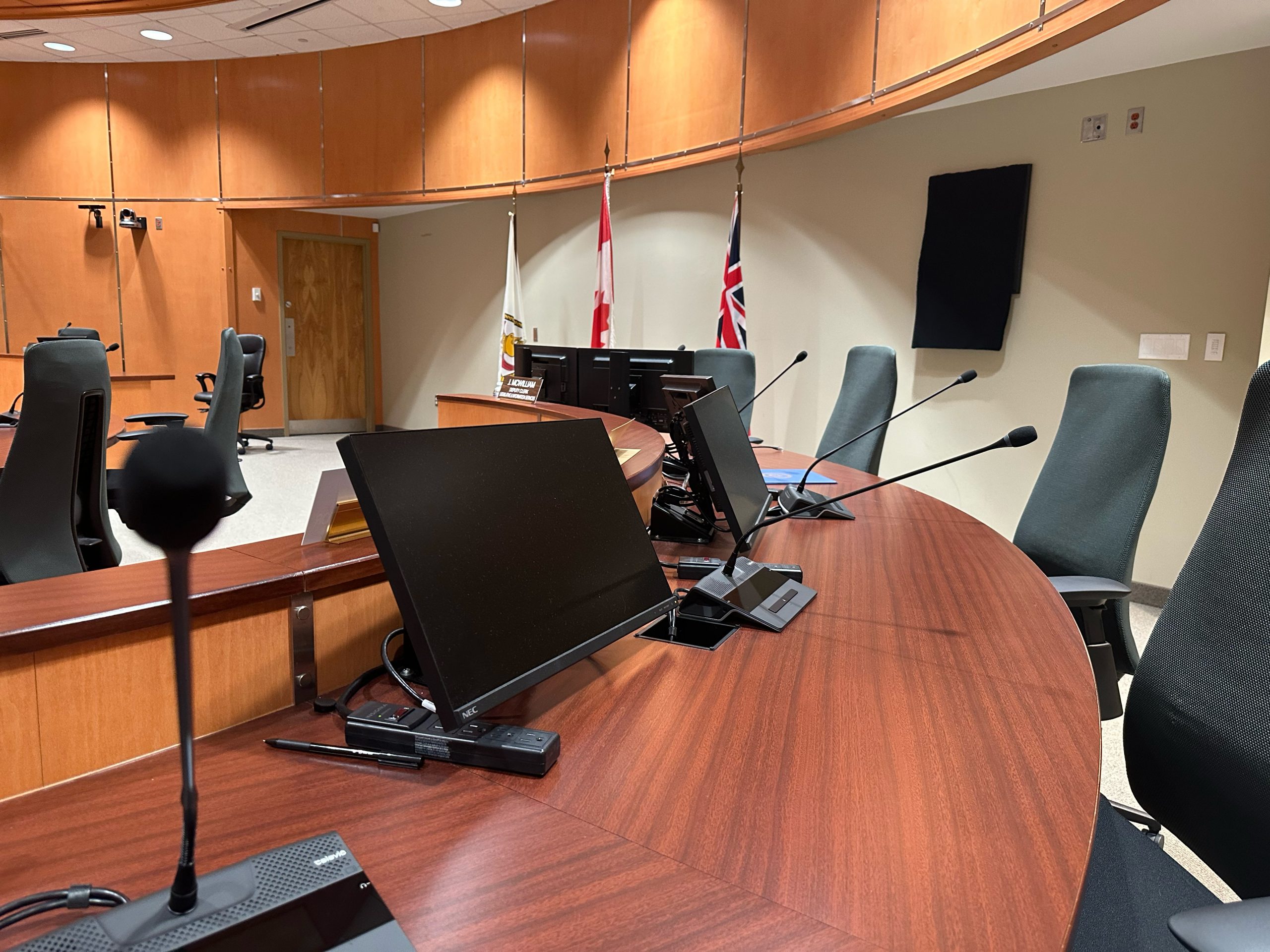 A row of empty chairs and microphones at a curved wooden desk in a modern meeting room with two flags in the background.