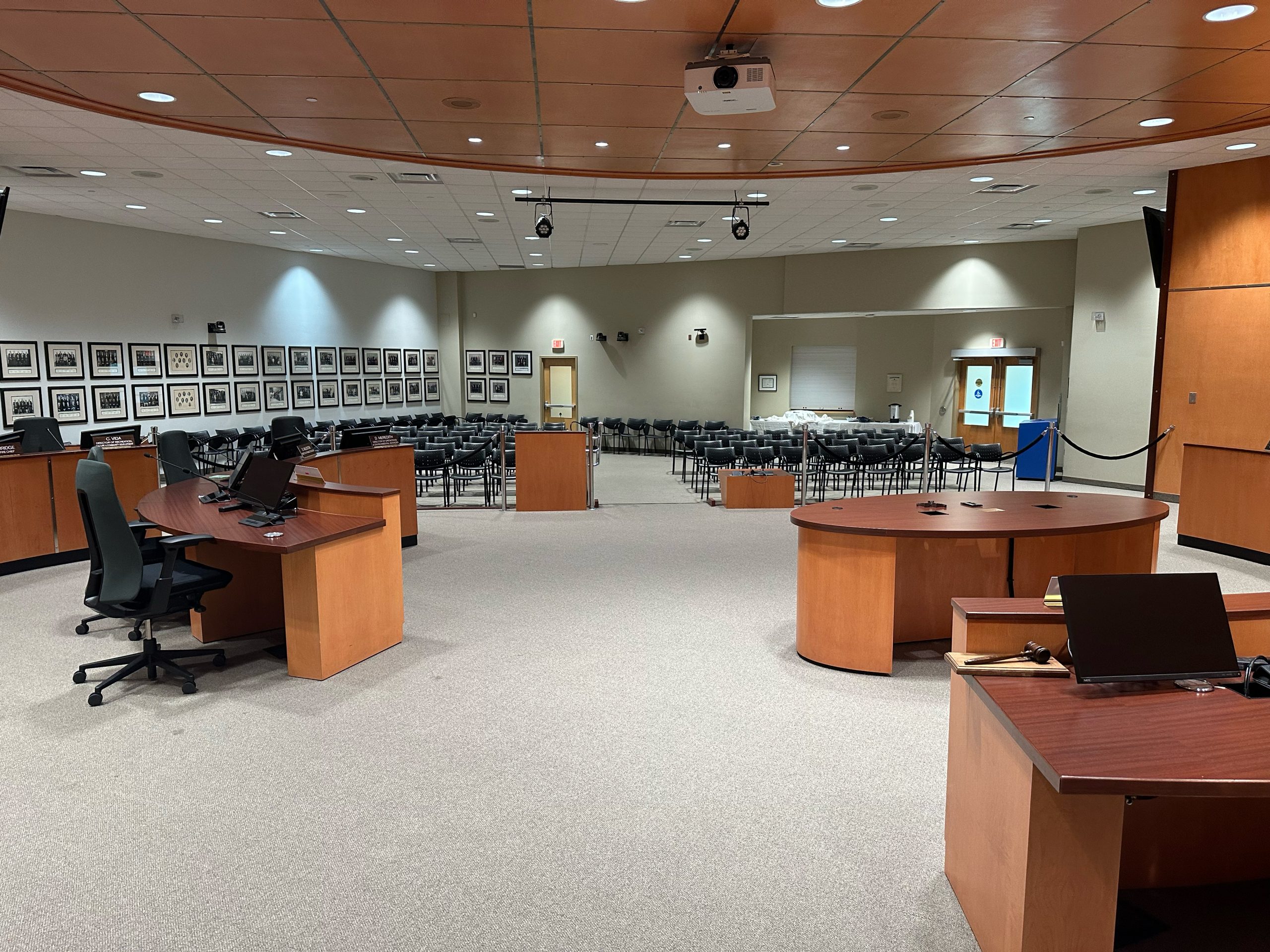 Empty council chamber with wooden desks, rows of chairs, framed photos on the wall, and a projector on the ceiling.