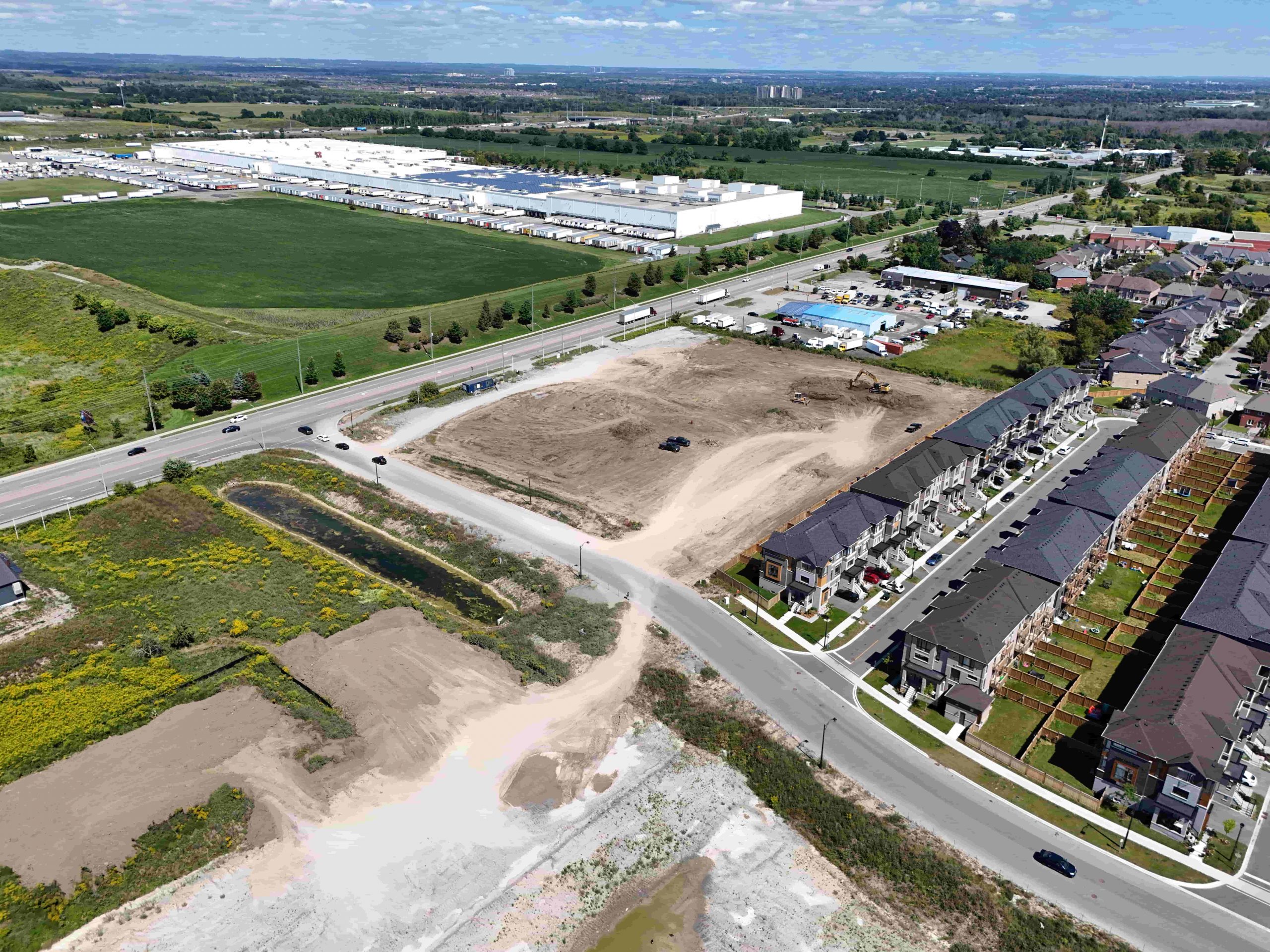 Aerial view of a construction site beside a residential neighbourhood and industrial buildings, with roads and green fields nearby.