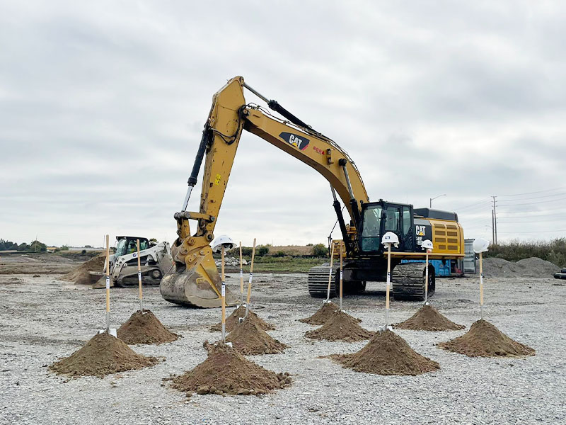 A yellow digger is parked near several small piles of earth marked with stakes on a gravelled lot.