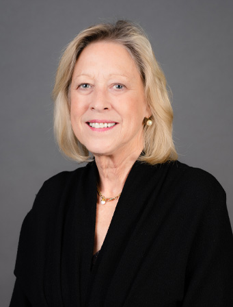 A middle-aged woman with blonde hair wearing a black top and gold necklace, smiling against a grey background.