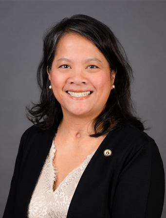 A woman with dark hair smiles at the camera, wearing a black blazer over a light blouse, in front of a grey background.