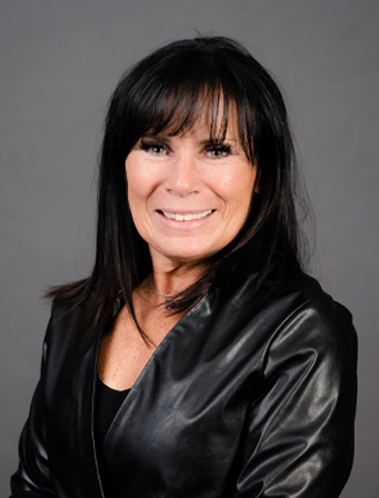 Woman with straight dark hair wearing a black jacket, smiling in front of a plain grey background.