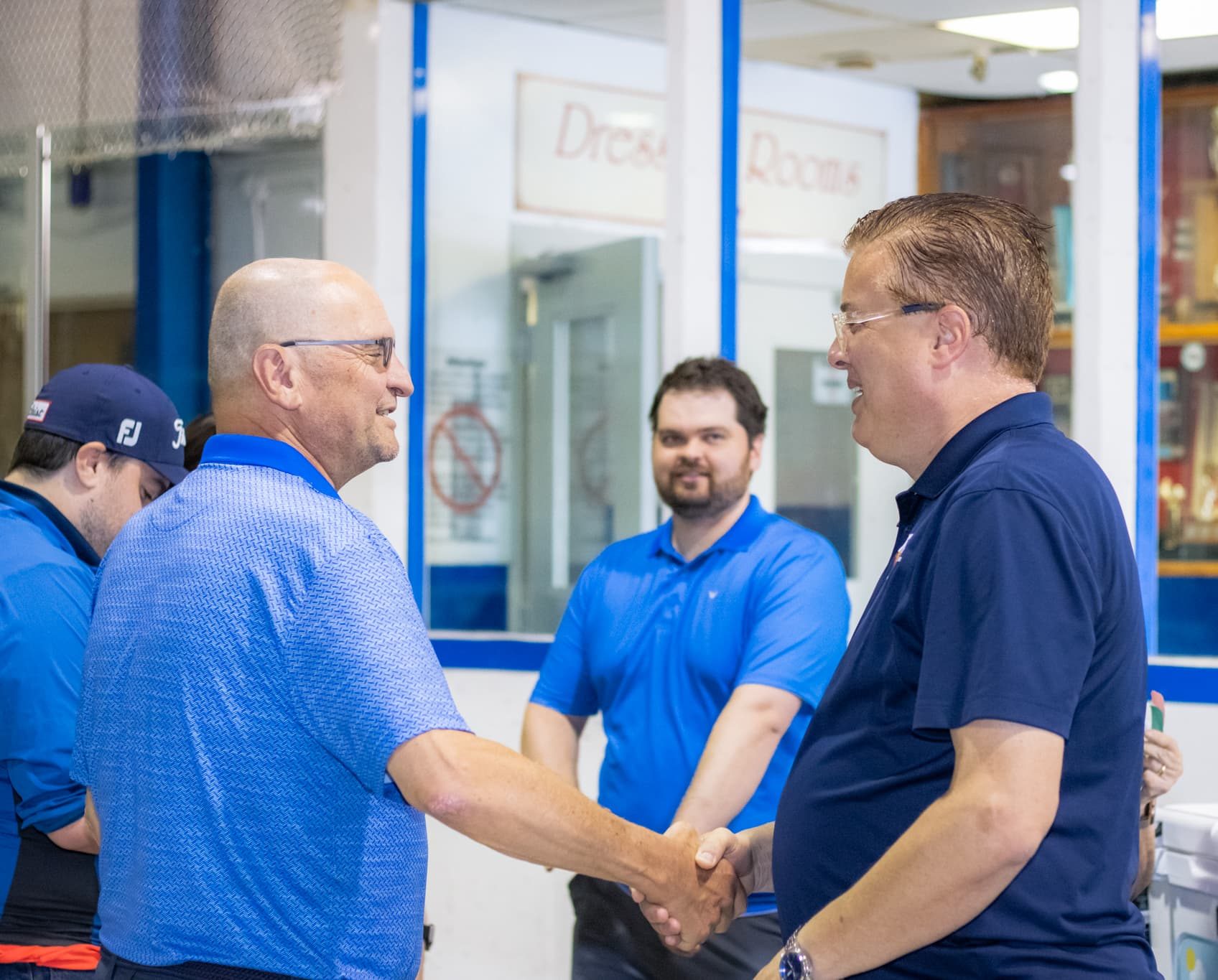 Two men in blue shirts shake hands in an indoor setting whilst others stand nearby.