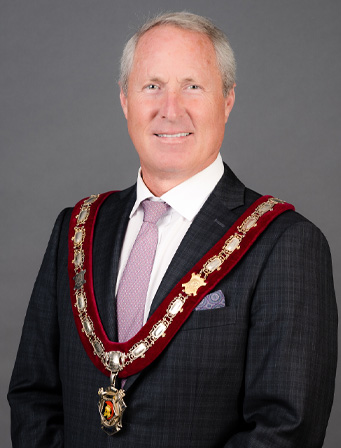 A man in a suit and tie wears a ceremonial chain of office and poses against a plain grey background.