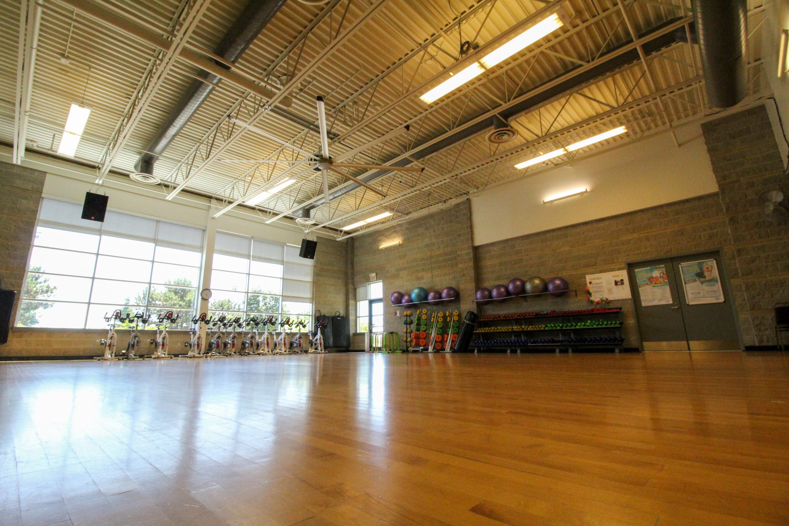 Empty fitness studio with wooden floor, exercise bikes by windows, and racks of weights and stability balls against the wall.
