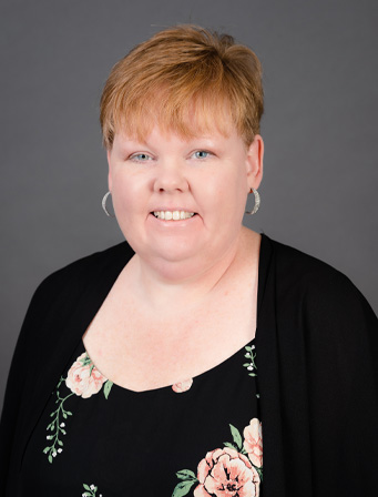 A woman with short light brown hair, wearing hoop earrings and a black floral top, smiles against a grey background.