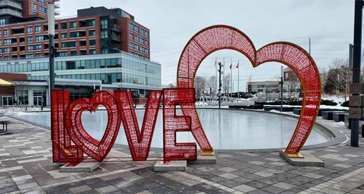 Large red "LOVE" sculpture with heart shapes stands beside an empty outdoor skating rink in an urban area.