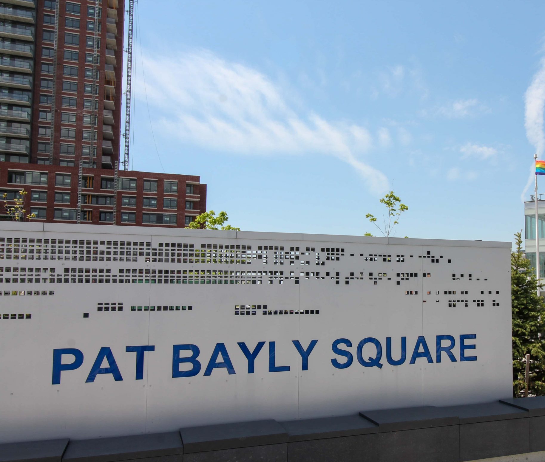 Sign reading "Pat Bayly Square" in front of modern buildings with flags and rental living advert in the background.
