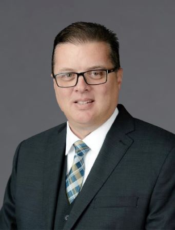 Man wearing glasses, a dark suit, white shirt, and checked tie, posing in front of a plain grey background.