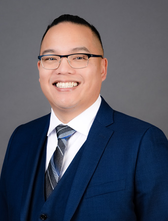 Smiling person wearing glasses, a blue suit, white shirt, and striped tie, posing against a plain grey background.