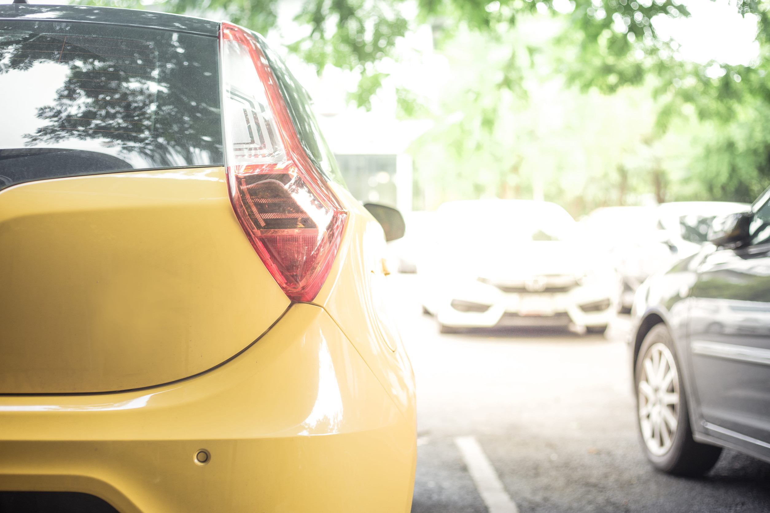 Close-up of the rear left side of a yellow car parked in a car park with other cars and trees in the background.
