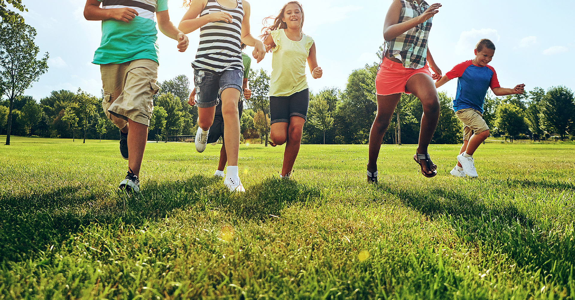Five children run across a grassy park field on a sunny day, trees in the background.