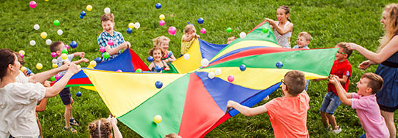 Children and adults playing with a colourful parachute and plastic balls on a grassy field.