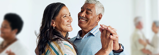 Smiling older couple dancing together indoors, with other pairs dancing in the blurred background.