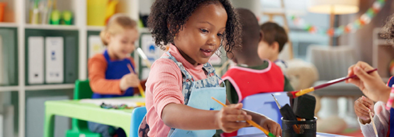 Young children sit at tables in a classroom, painting and engaging in arts and crafts activities.
