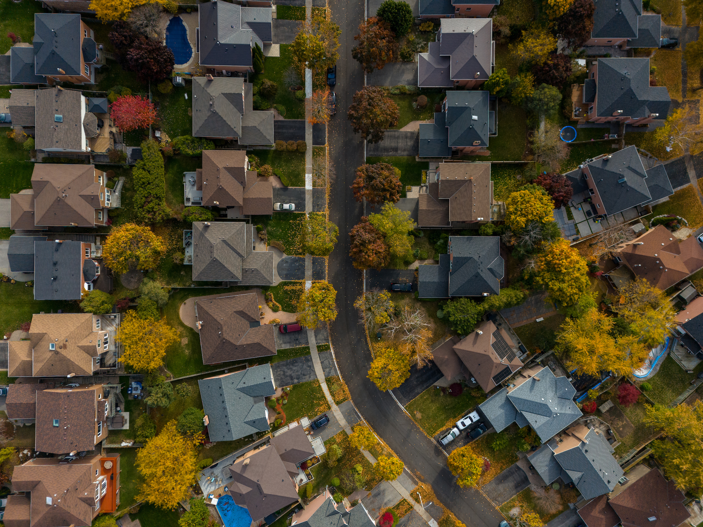Aerial view of a suburban neighbourhood with houses, trees with autumn foliage, and cars parked along the street.