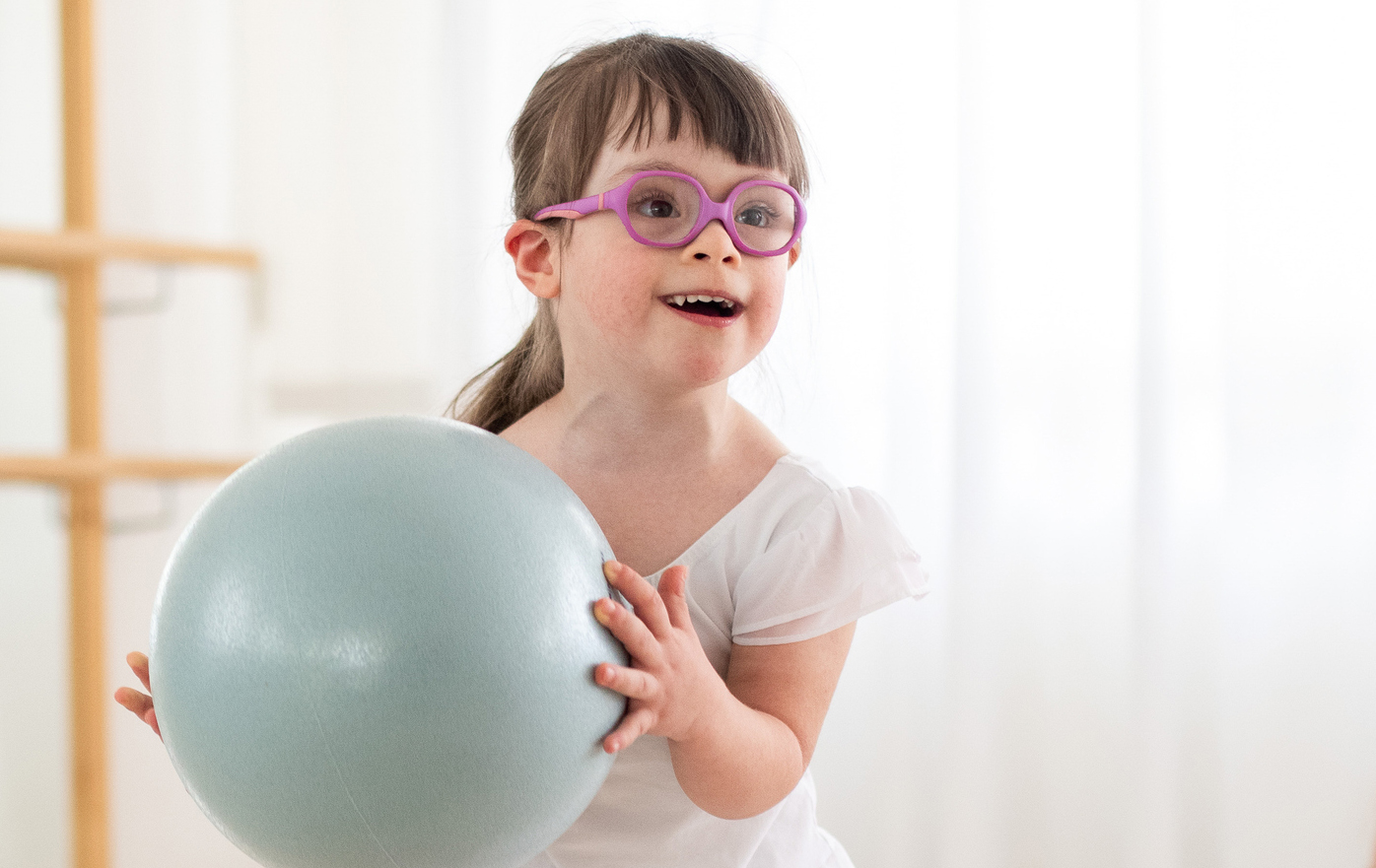 Girl with glasses holding an exercise ball in a bright room, smiling and looking forward.